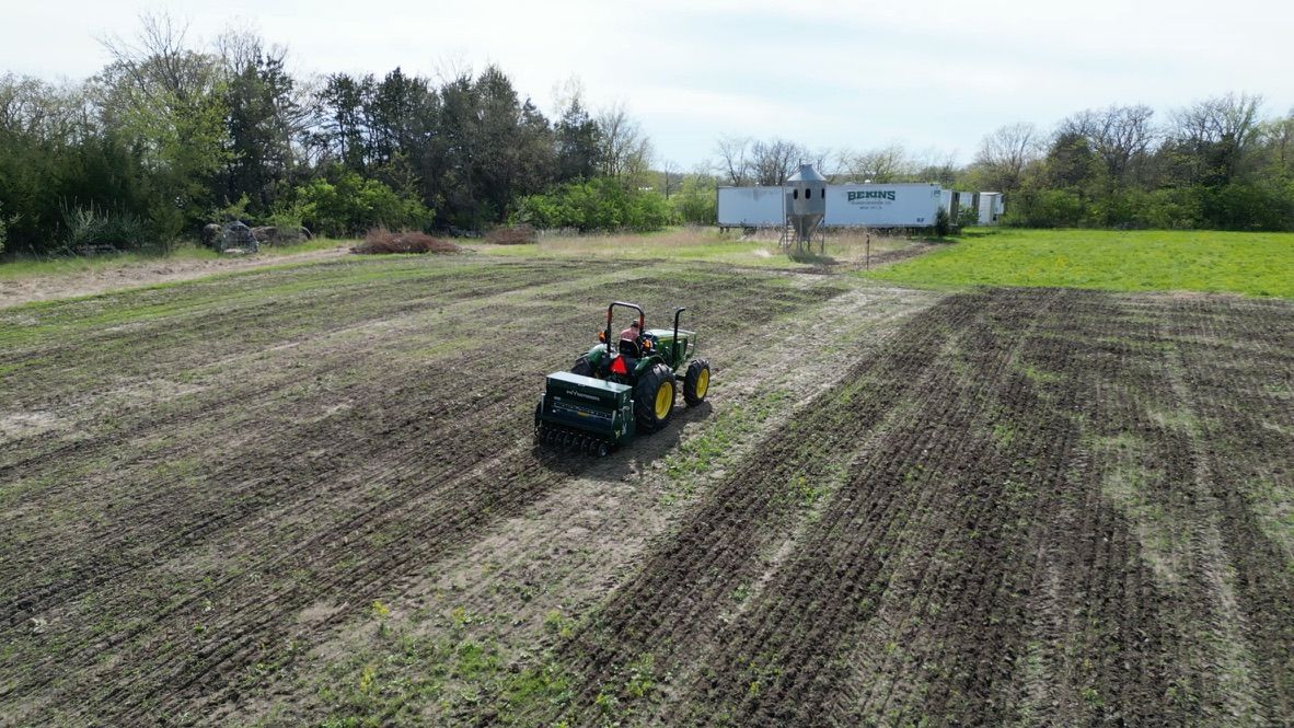 Small tractor towing a planter across a plowed field near a farmhouse and trees