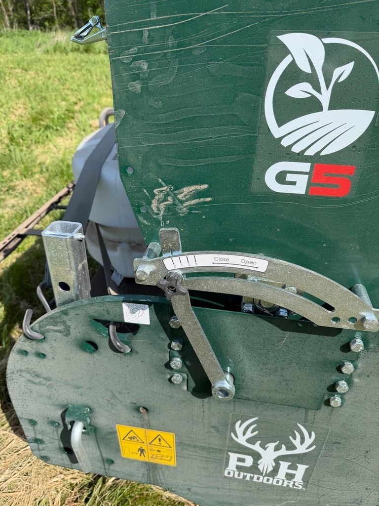 Green farm machine with deer logo, visible metal wheel and yellow warning sticker outdoors in grass