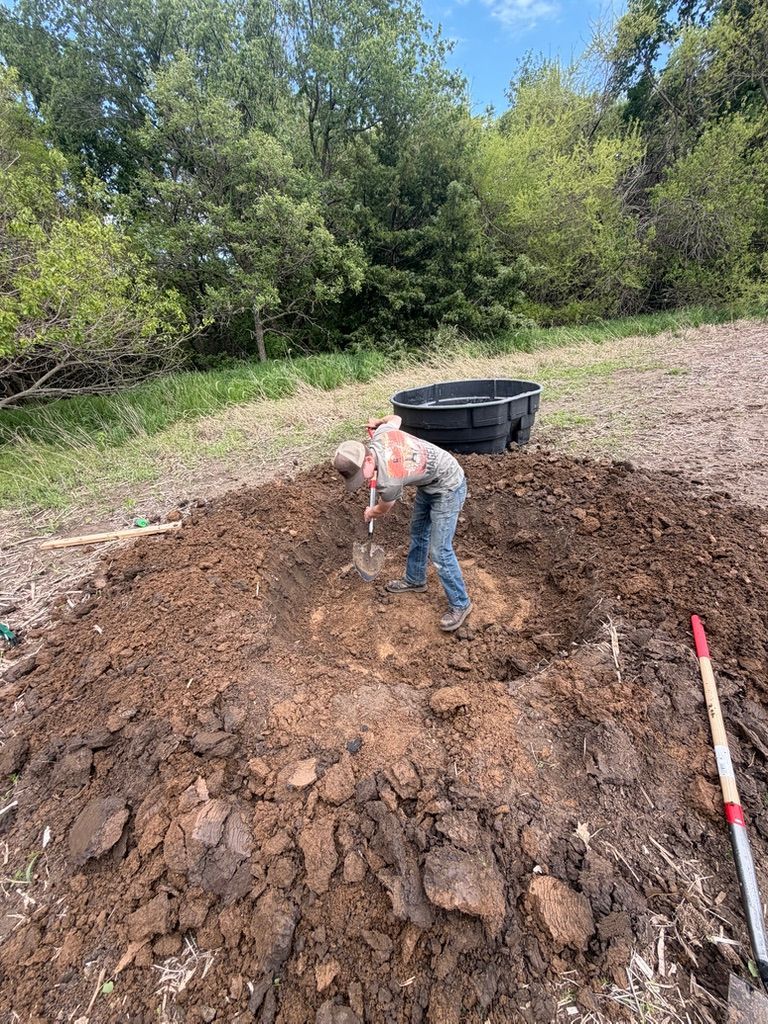 Person shoveling dirt beside a black bin in a muddy outdoor area with trees in the background