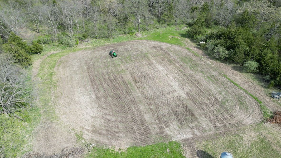 Aerial view of a freshly tilled field bordered by trees, with a small green tractor near the center.