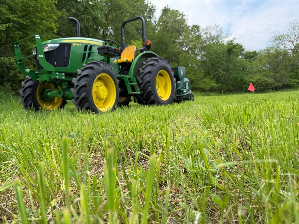 Green tractor with yellow wheels working in a grassy field under a cloudy sky