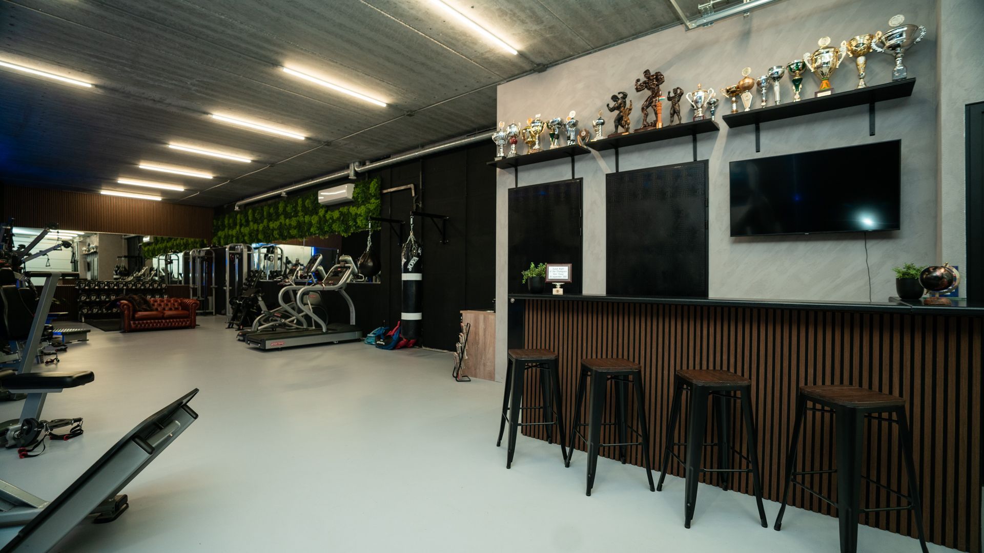 Modern fitnesscentrum met gewichtsbanken, hometrainers en een lange houten receptiebalie met trofeeën erboven.
