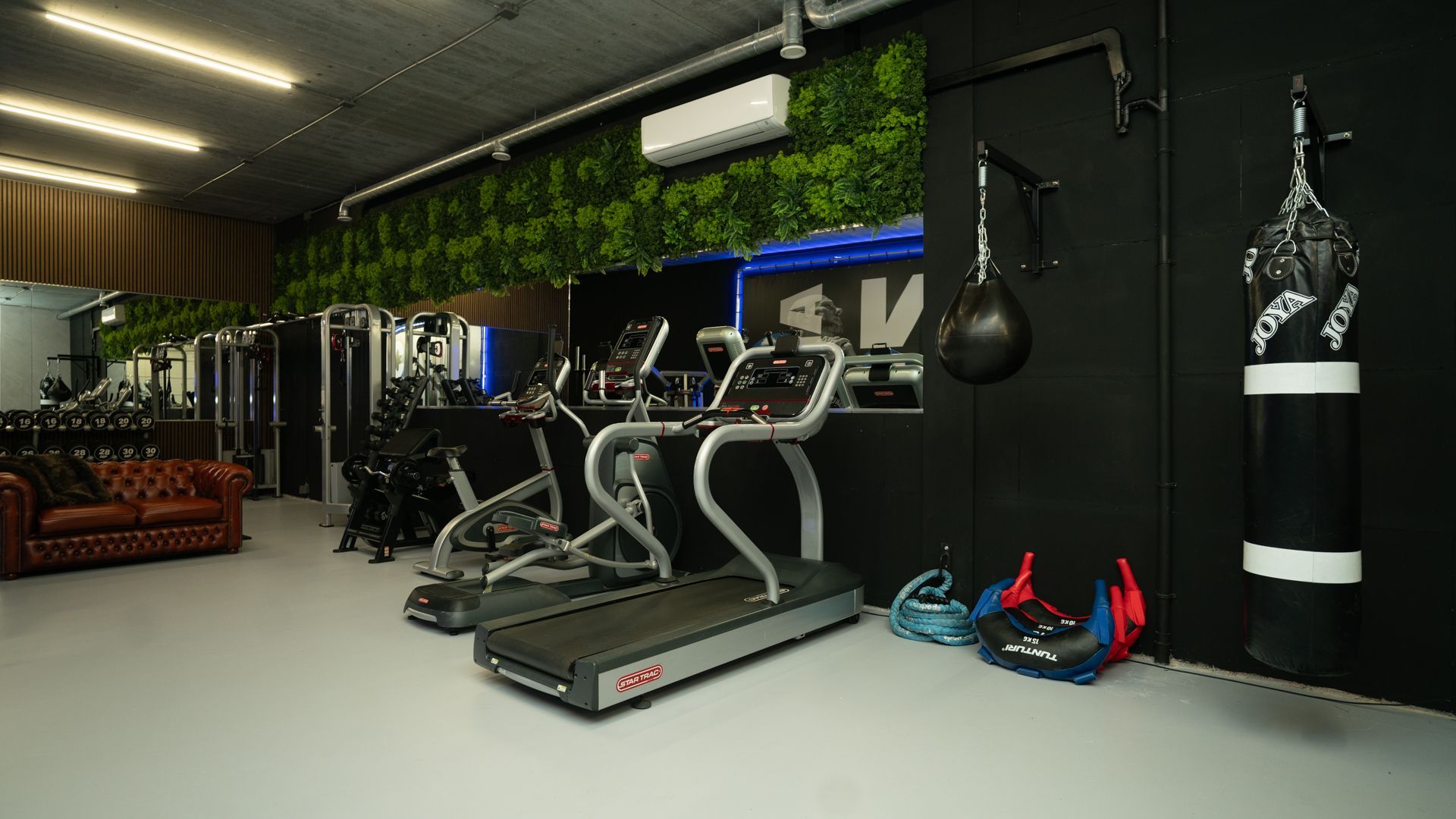 Moderne sportschool met loopbanden, een hangende boksbal en blauw-rode fitnessapparatuur tegen een zwarte muur.