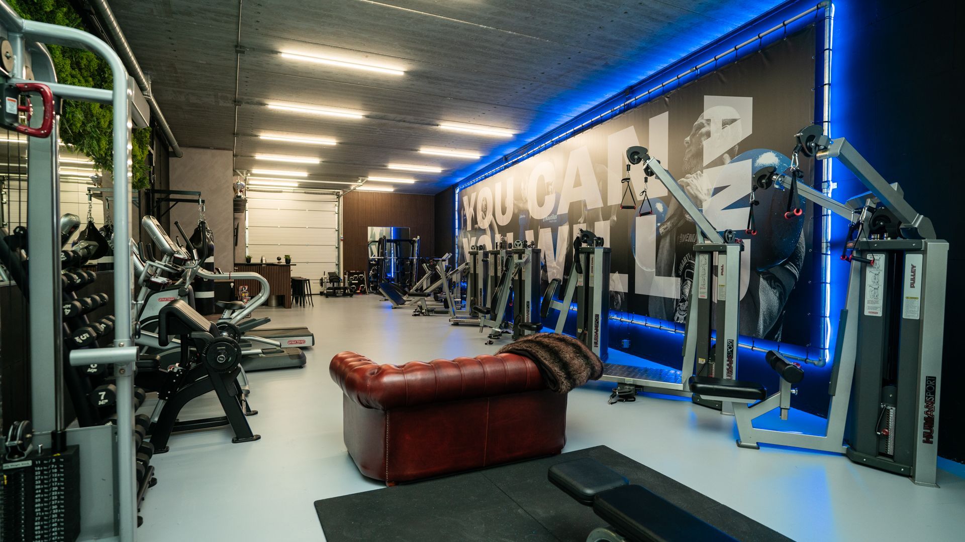 Modern fitnesscentrum met gewichtsapparaten, banken en blauwe accentverlichting.