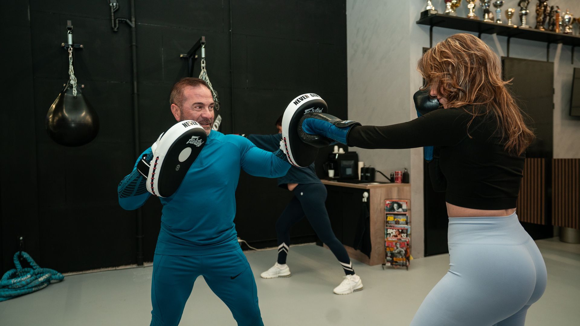 Mensen trainen met bokspads in een sportschool, waarbij ze middentrappen en stoten oefenen.