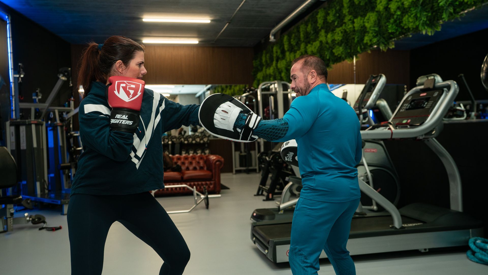 Twee mensen boksen in een sportschool, een met een rode hoofdbeschermer tegenover een trainer in een blauw pak.