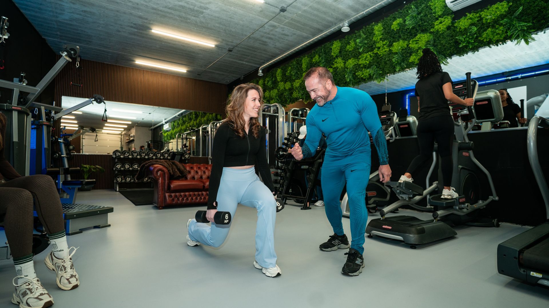 Twee mensen trainen in een sportschool; de ene doet lunges terwijl de andere in de buurt coacht.