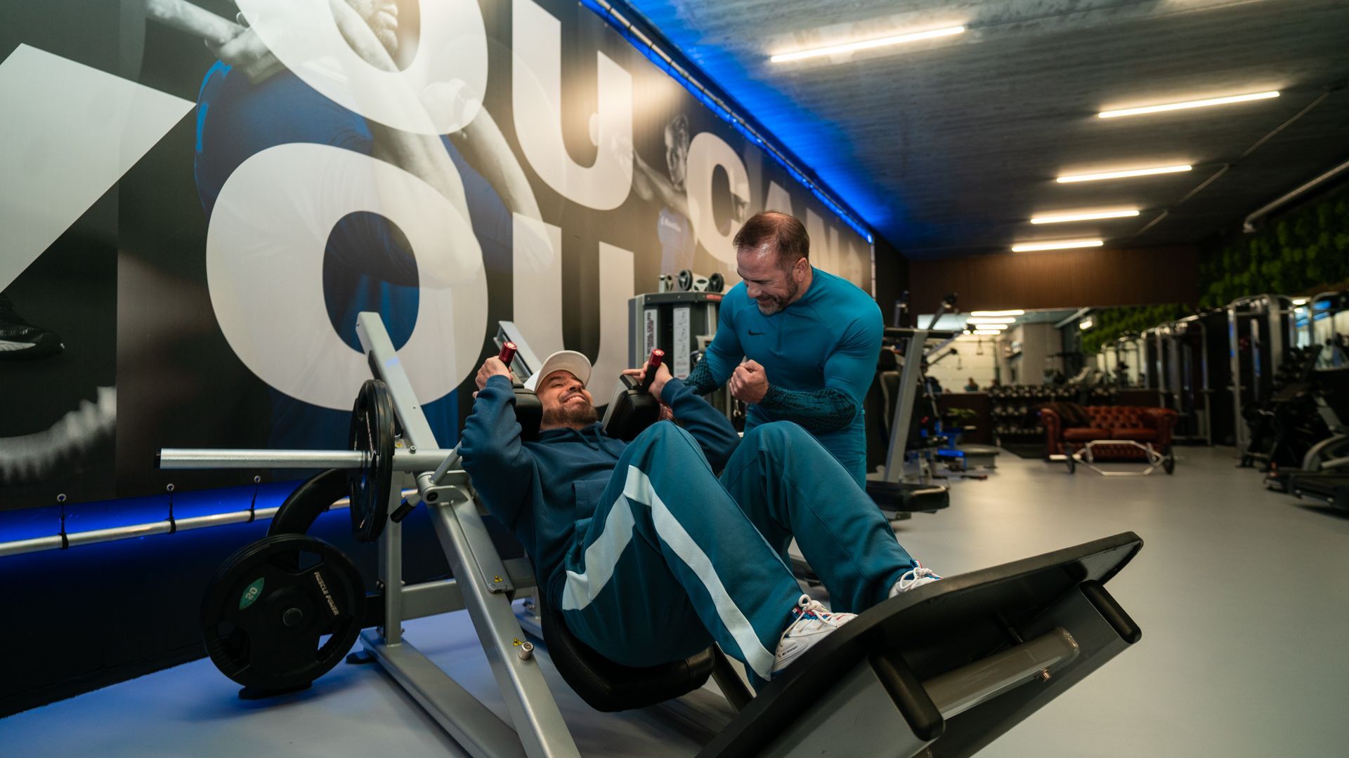 Twee mensen trainen op een legpress-apparaat in een sportschool, met blauwe apparatuur en fel plafondlicht.