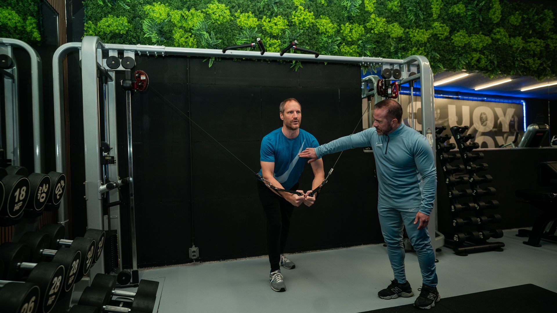 Twee mannen demonstreren in een sportschool een rek- of overdrachtsoefening in de buurt van een kabelmachine en gewichten.