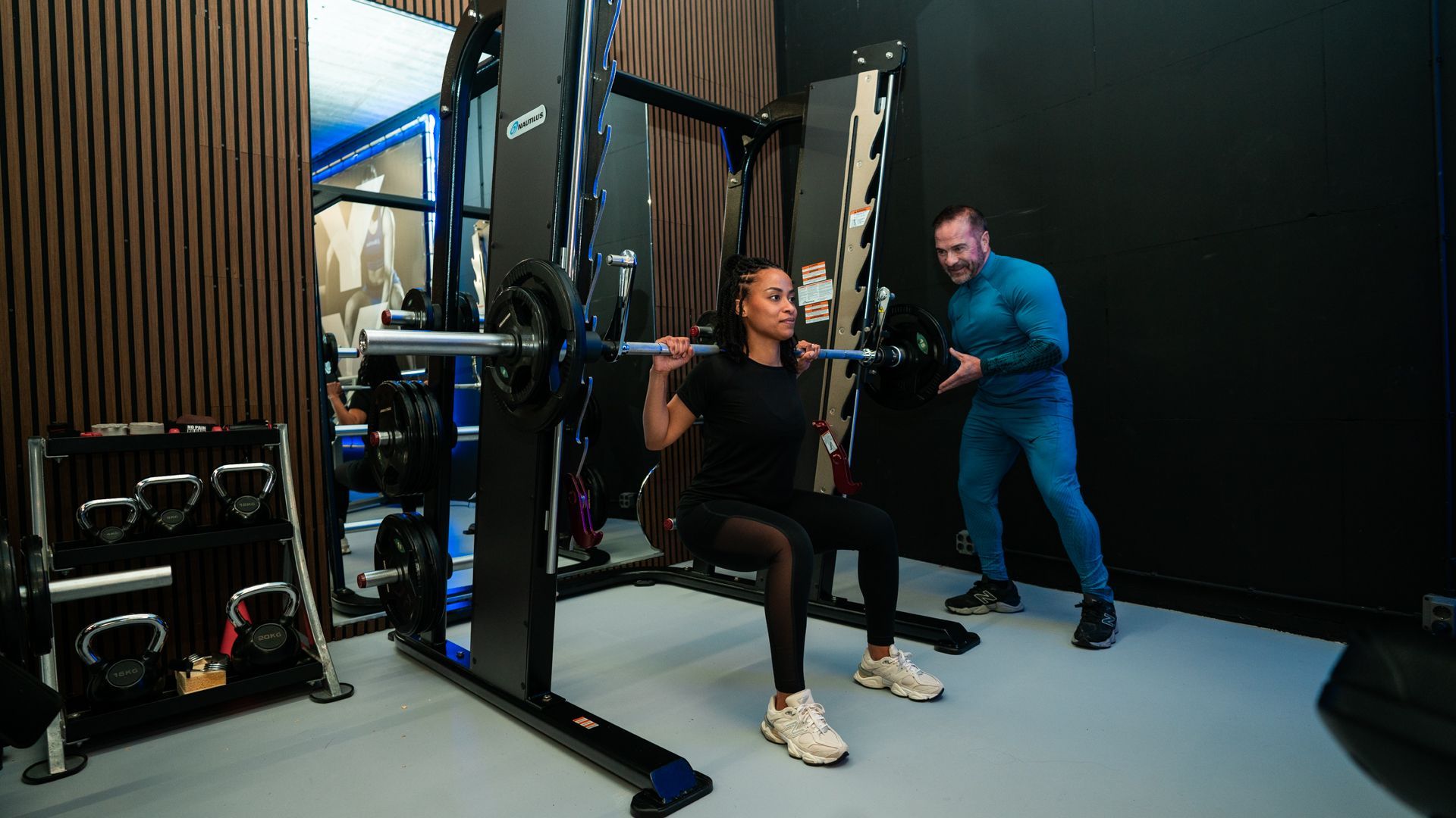 Een persoon die oefeningen doet op een kabelmachine, terwijl een trainer assisteert in een sportschool.