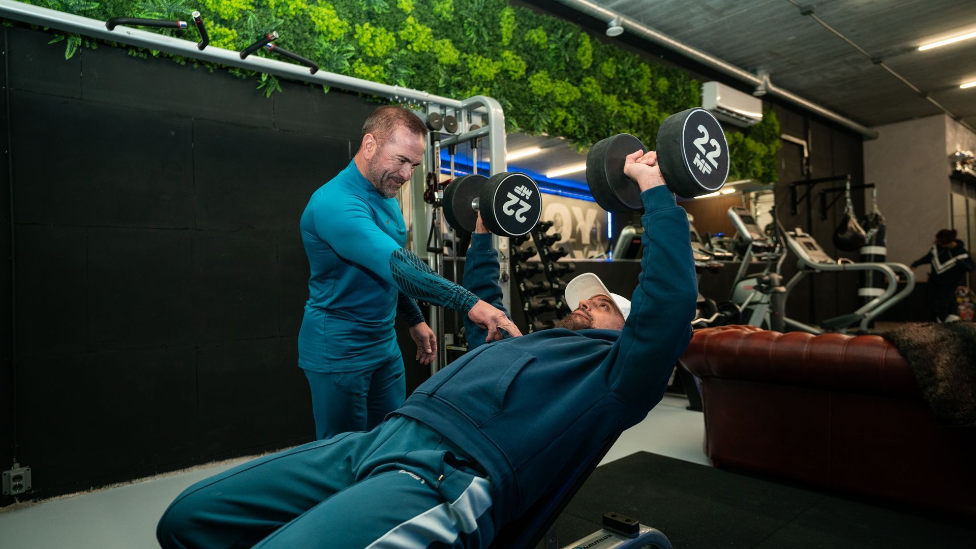 Een trainer helpt iemand met het tillen van dumbbells in een fitnessruimte.