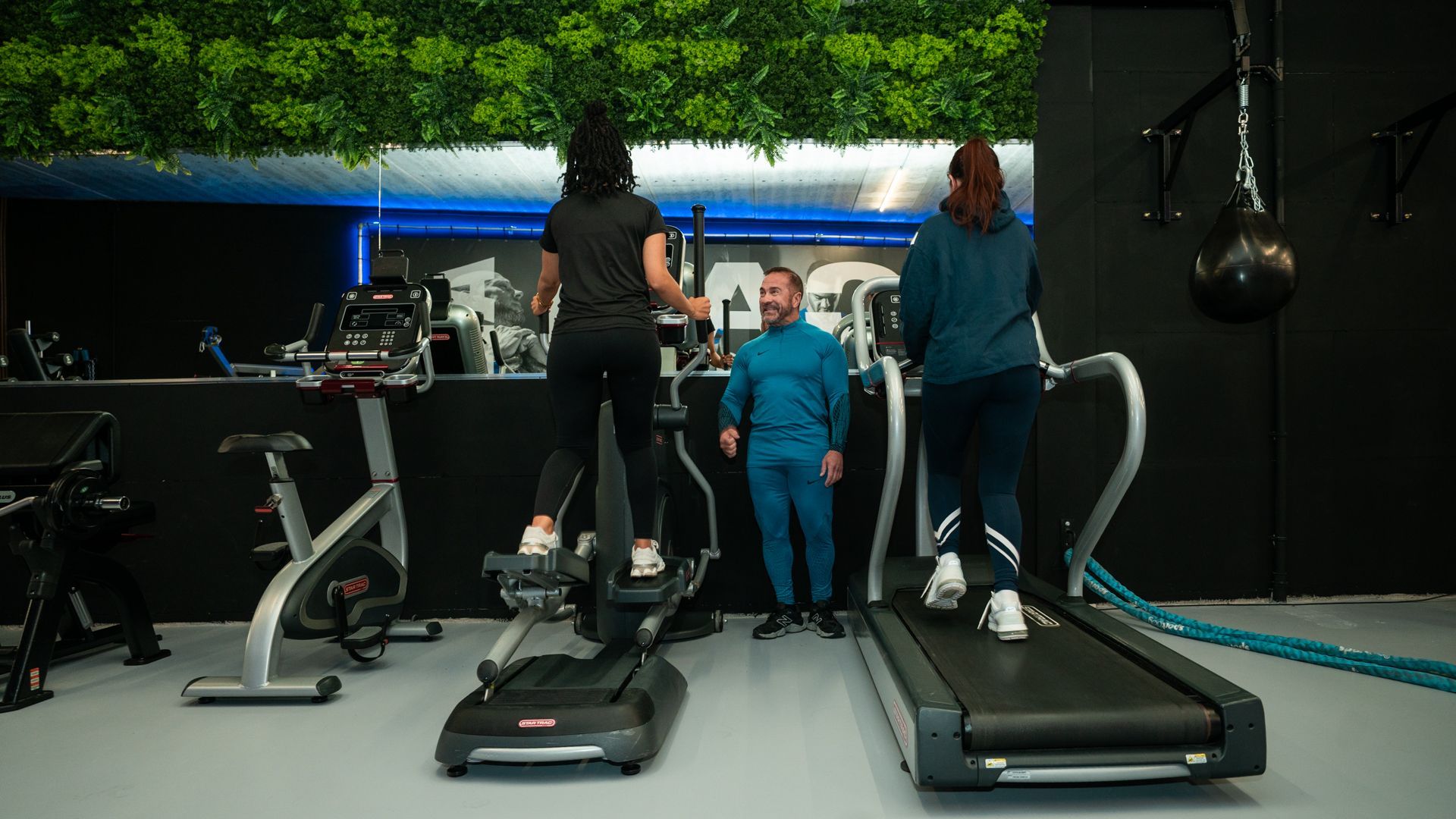 Mensen die sporten op loopbanden in een sportschool, met groen als decoratie en fitnessapparatuur op de achtergrond.