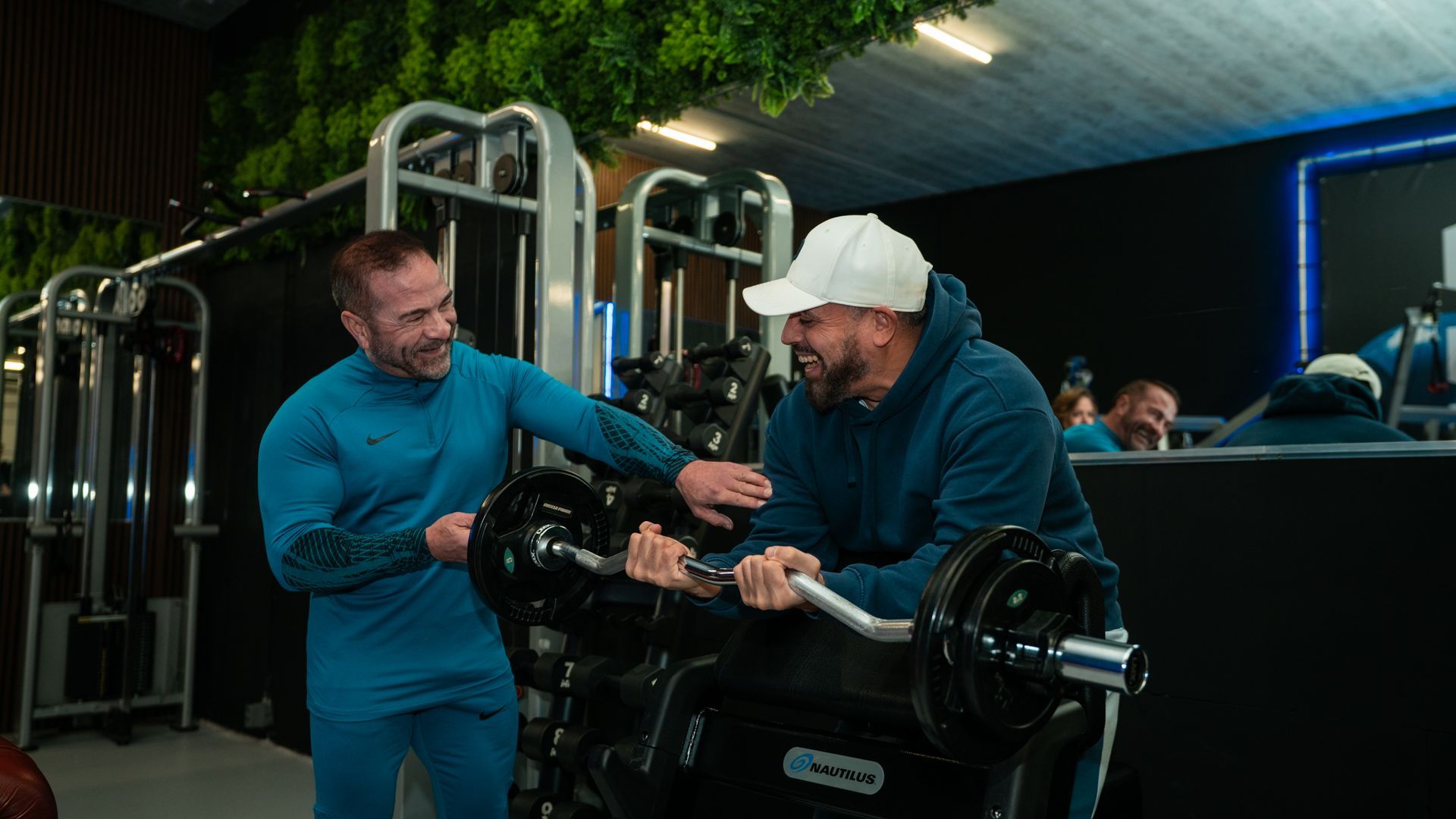 Twee mannen in turquoise sportkleding stellen een gewichtsapparaat af in een sportschool.