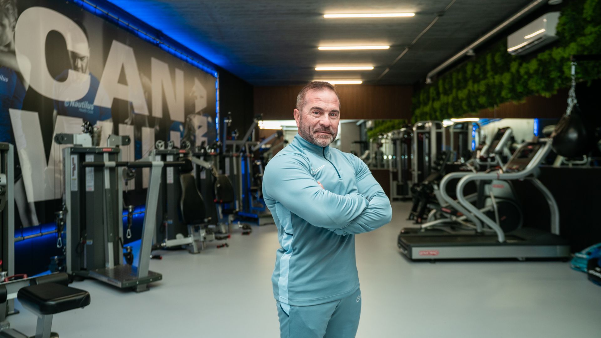 Een man staat met zijn armen over elkaar in een sportschool, met cardioapparatuur en de letters