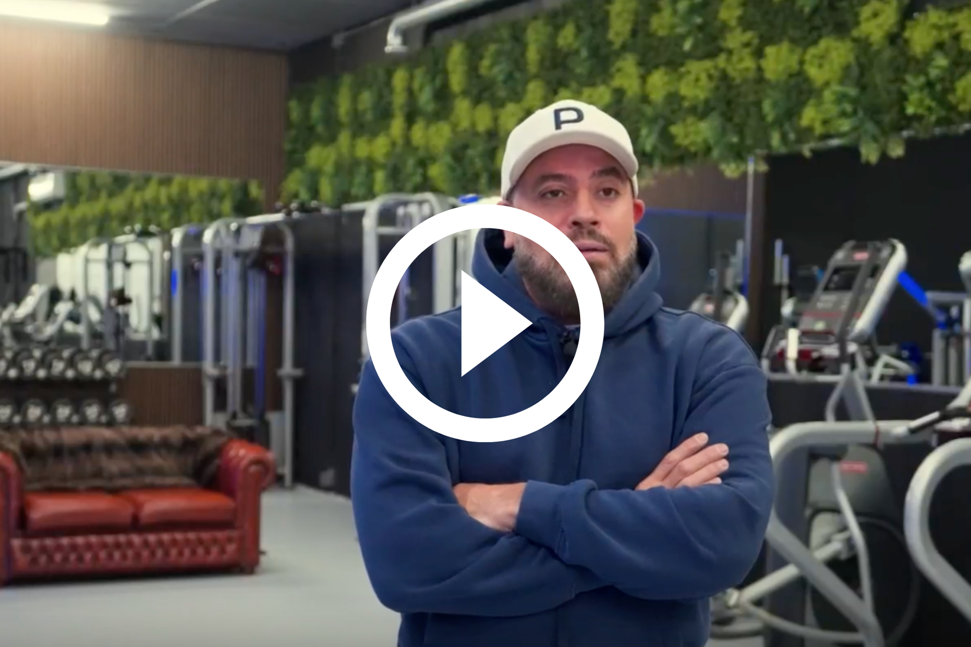 Een man staat met zijn armen over elkaar in een sportschool, met fitnessapparaten en een leren bank achter hem.