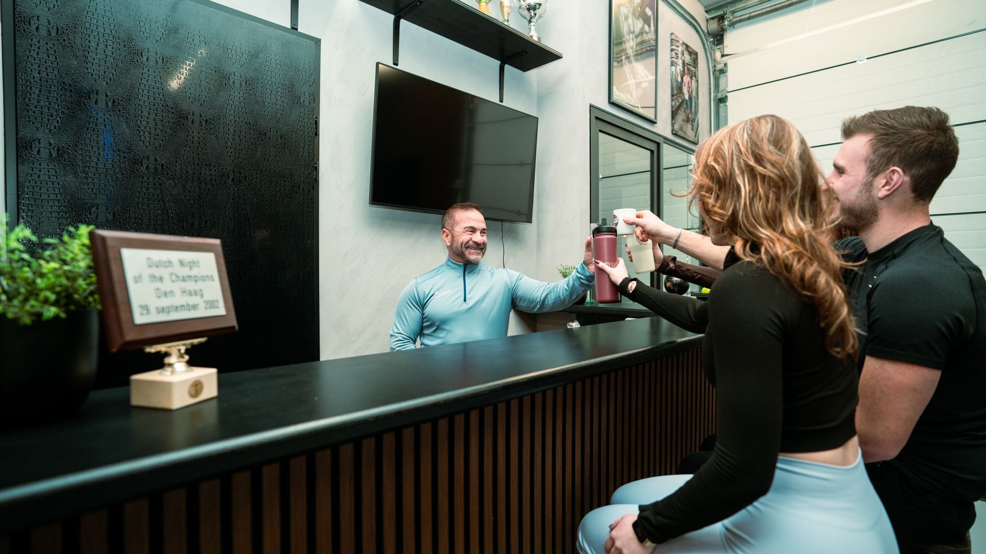 Mensen staan te praten aan een bar, met een barman erachter en een klein bordje op de toonbank.