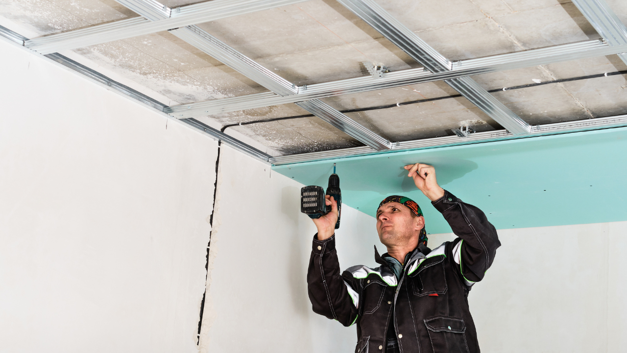 Man installing drywall on a ceiling using a drill, inside a room.