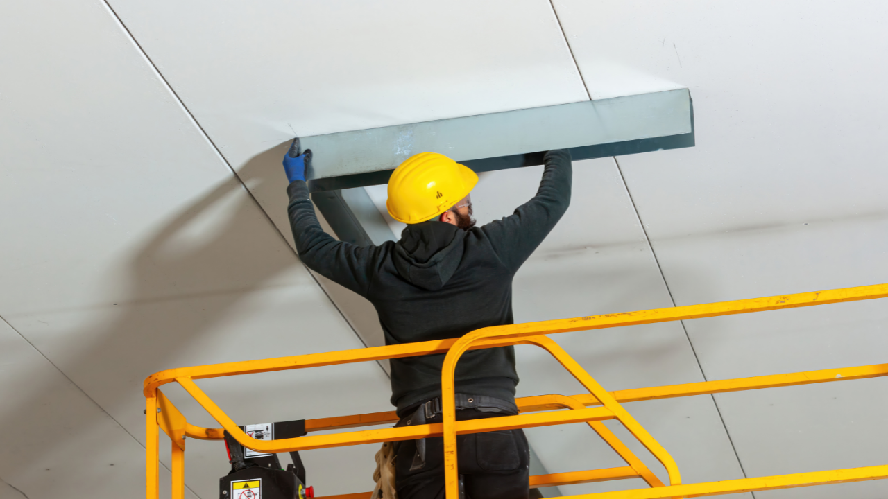 Construction worker on lift installs metal trim on ceiling, wearing a hard hat and gloves.
