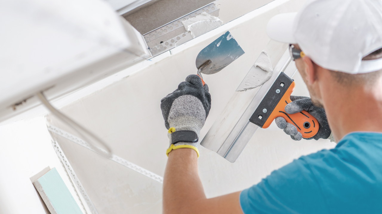 Person in a white cap and gloves applying drywall compound to a ceiling with a trowel.