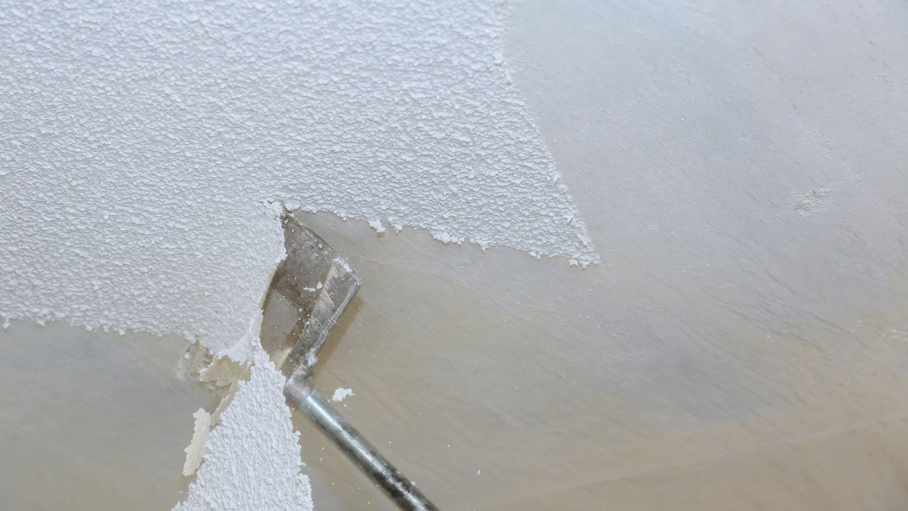A putty knife peeling off white, textured material from a neutral-colored surface.
