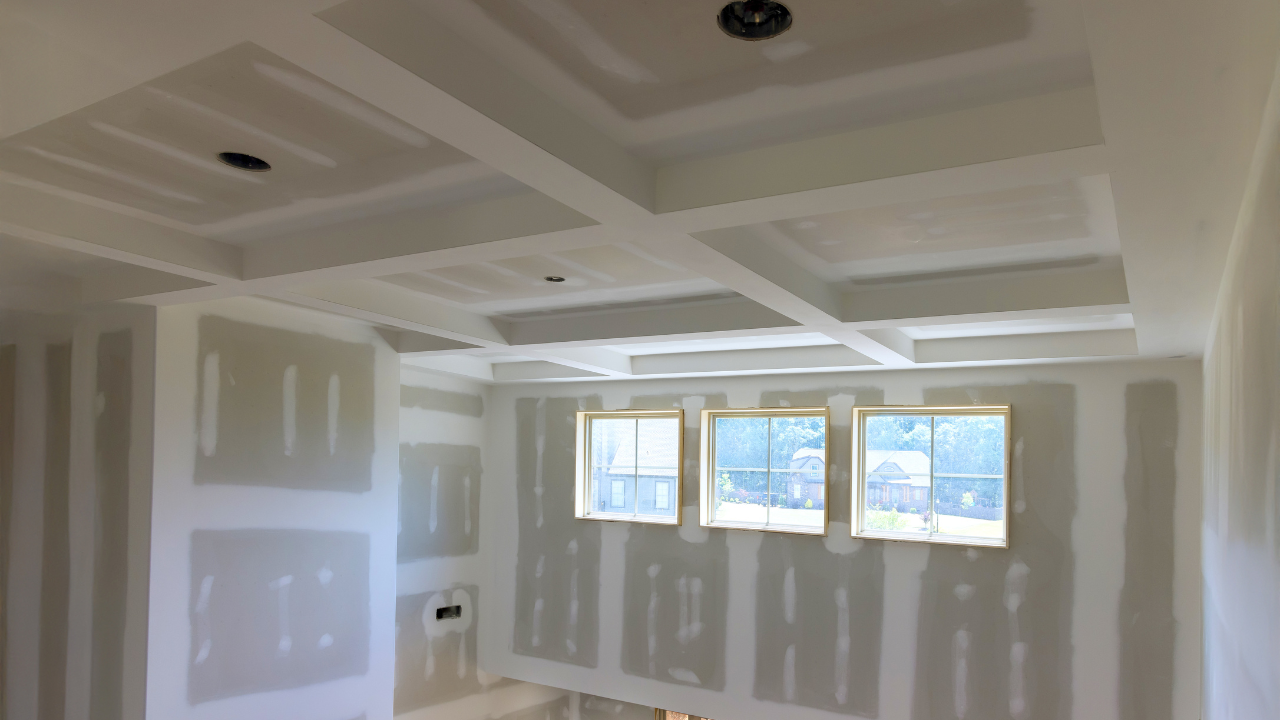Unfinished interior of a room, drywall walls and ceiling with rectangular beam design. Three windows are in the background.