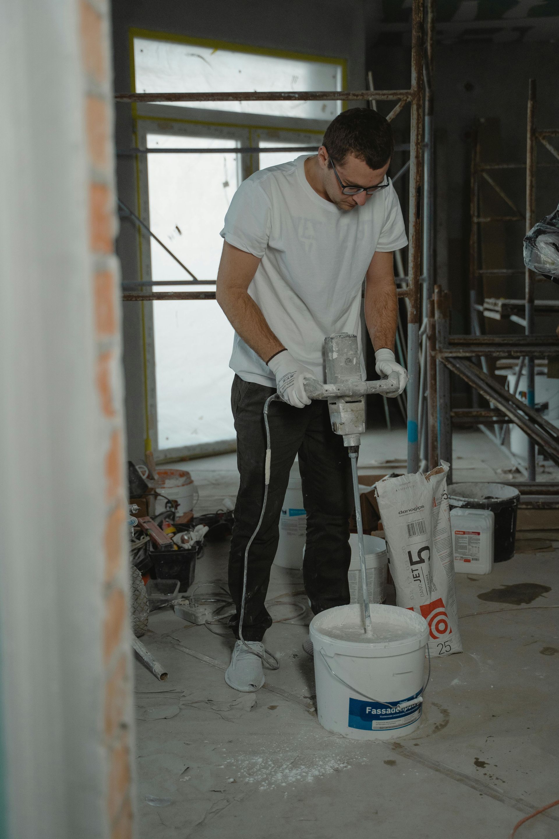 Man in white shirt mixing plaster in a bucket with a power mixer at a construction site.