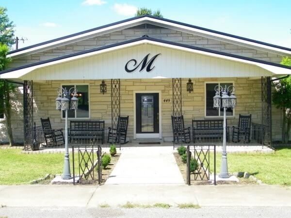 A single-story stone building with a front porch, iron accents, two lamp posts, and a large letter M on the gable.