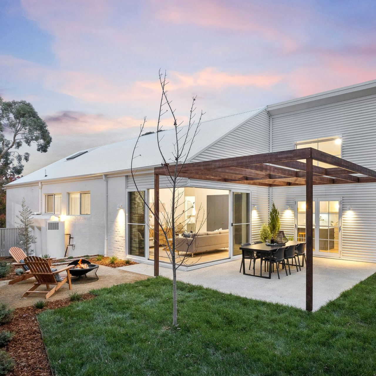 Modern white house with patio, pergola dining area, lawn, and warm lights at sunset