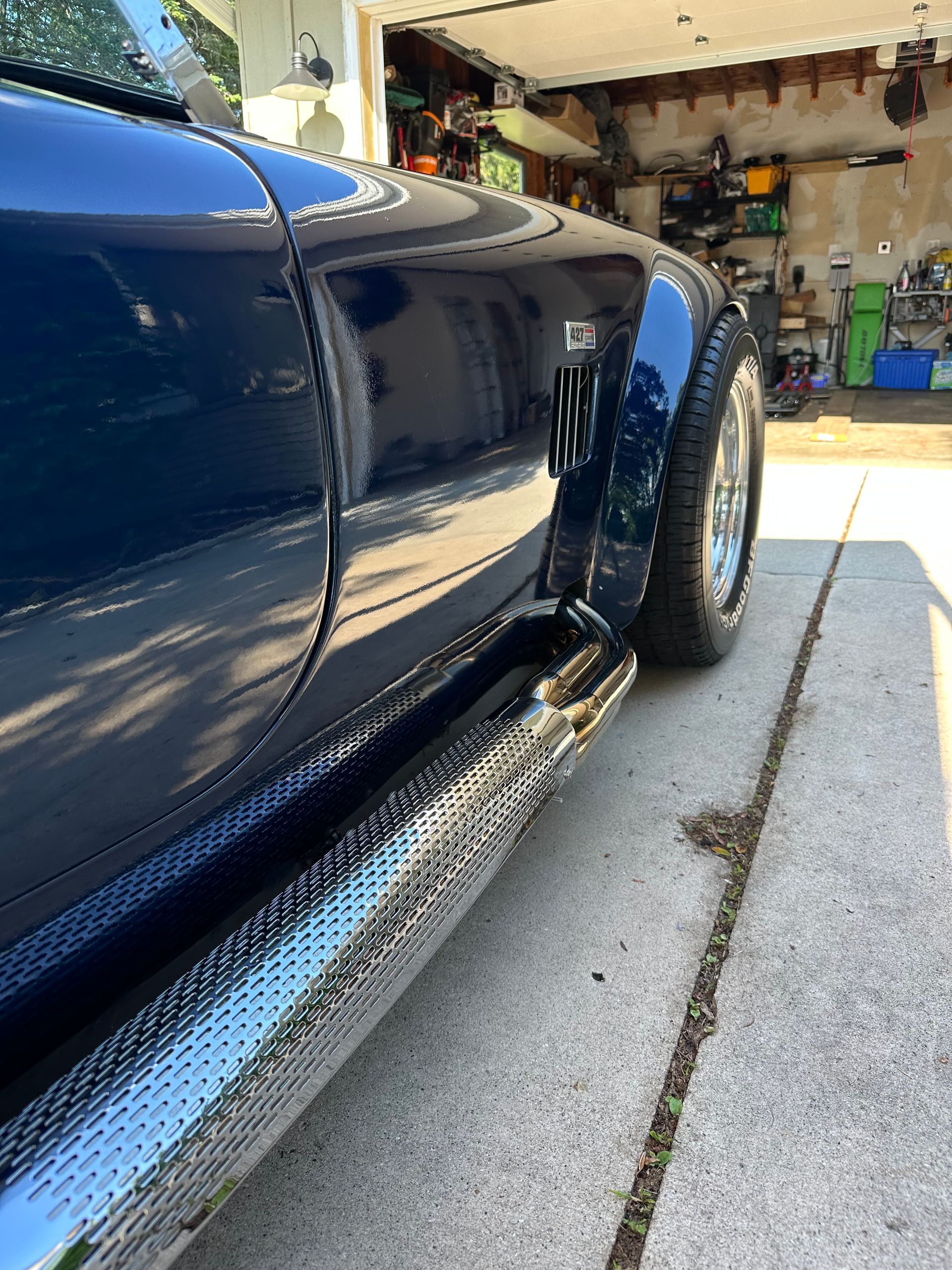 Blue sports car side panel and rear wheel in a garage driveway, with sunlight reflecting on the paint.