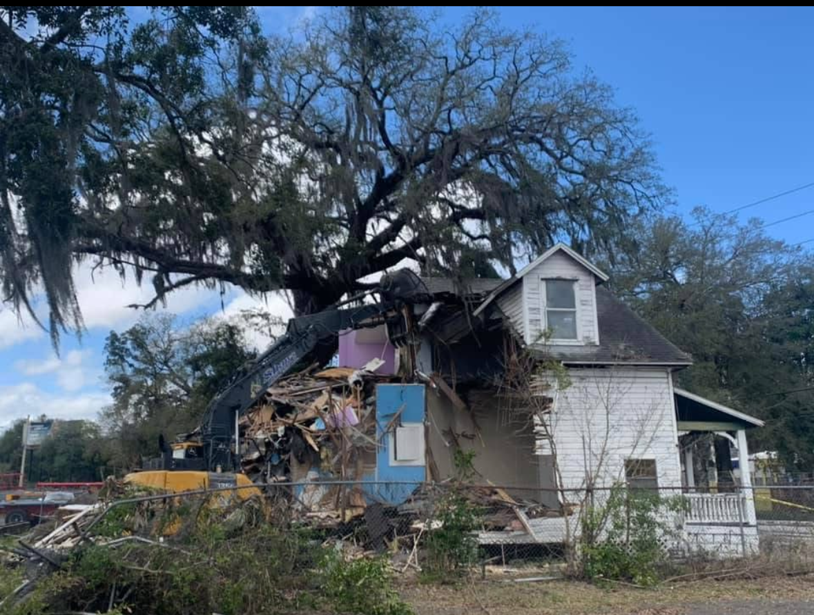 A damaged white house with a collapsed roof and debris, beneath a large tree with Spanish moss.
