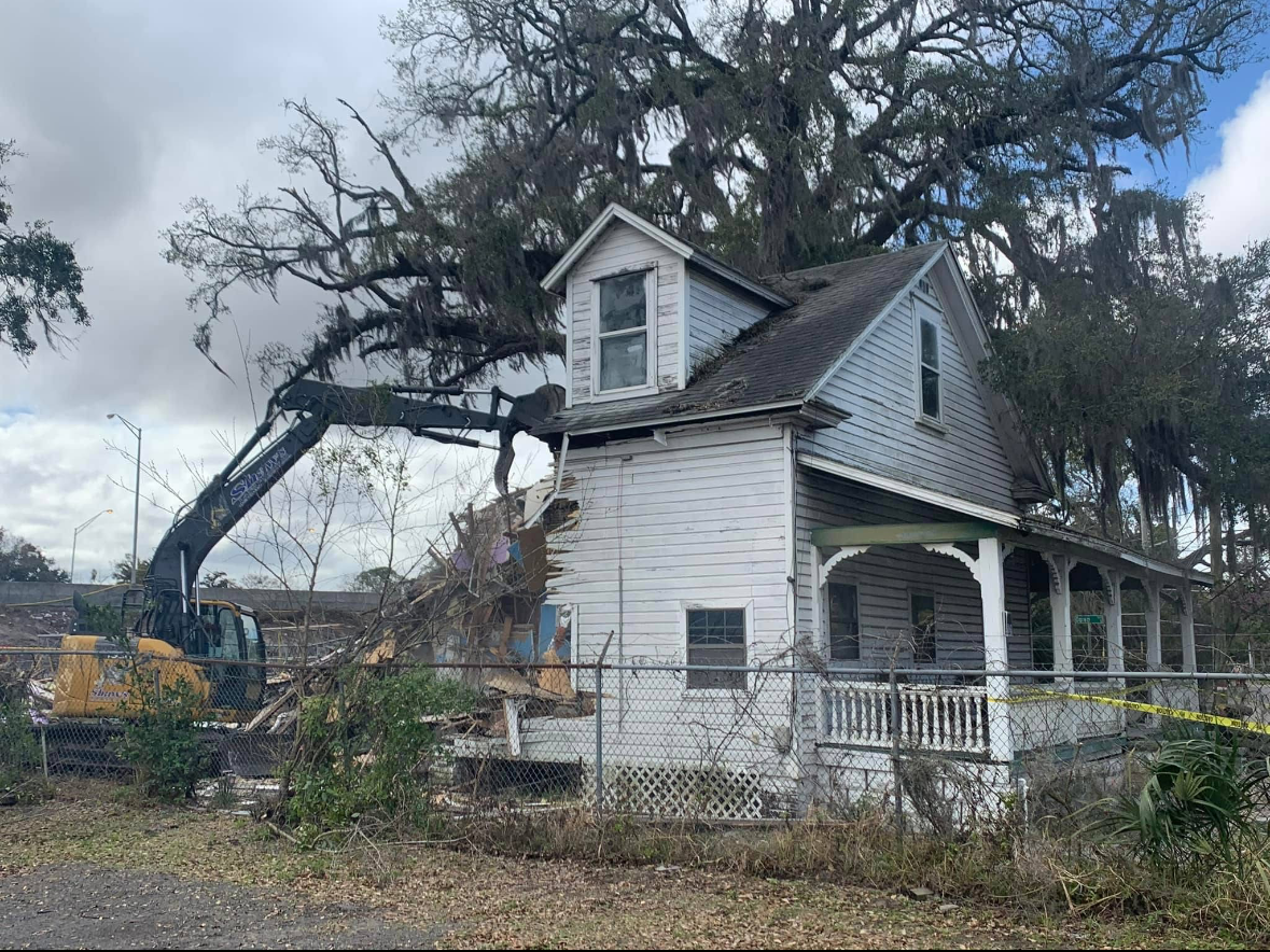 A house being demolished by an excavator. The house is white and blue, with a porch.