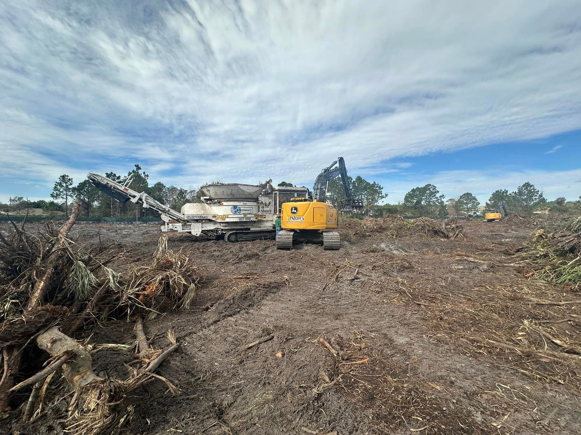 Heavy machinery clearing land, wood chipper and excavator visible. Cloudy sky overhead. Heavy machinery clearing land, wood chipper and excavator visible. Cloudy sky overhead.
