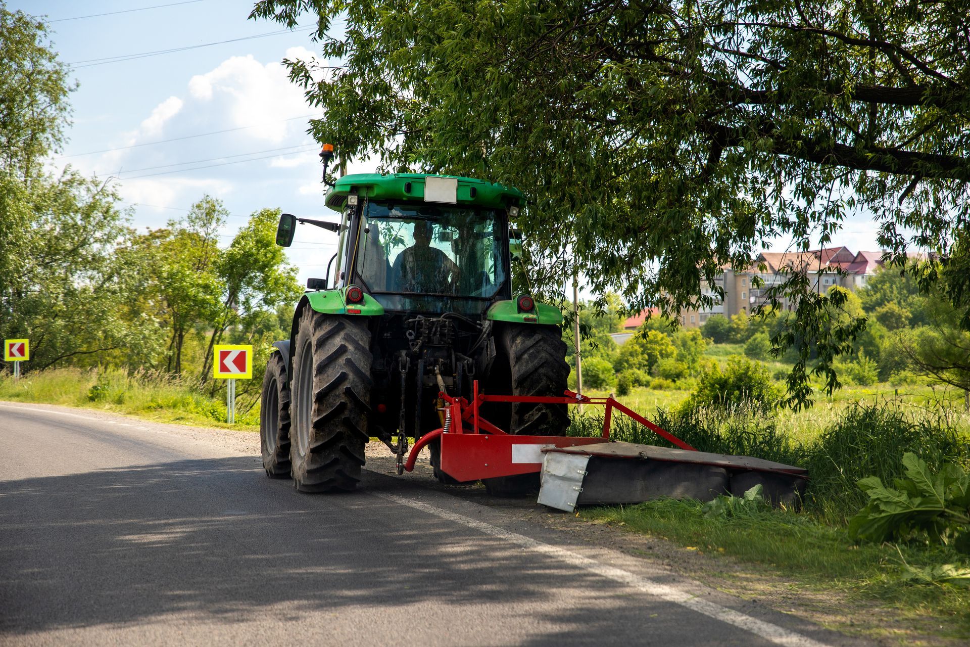 Green tractor mowing roadside grass beside a paved road under trees