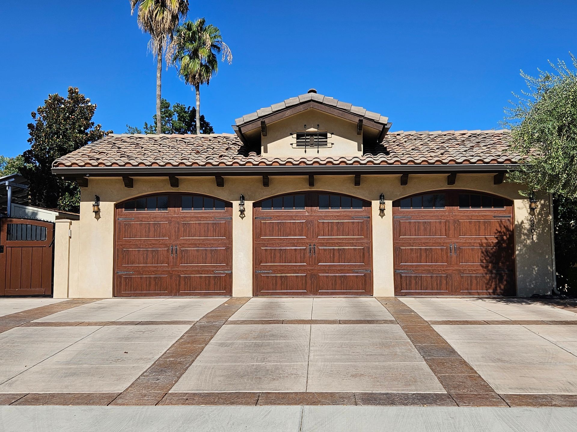 Three-car garage with brown doors and tan stucco exterior. Clay tile roof with a small dormer, blue sky, and palm trees.