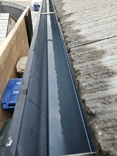 Water-filled black rain gutter along a concrete roof edge. Blue trash bins are visible below.