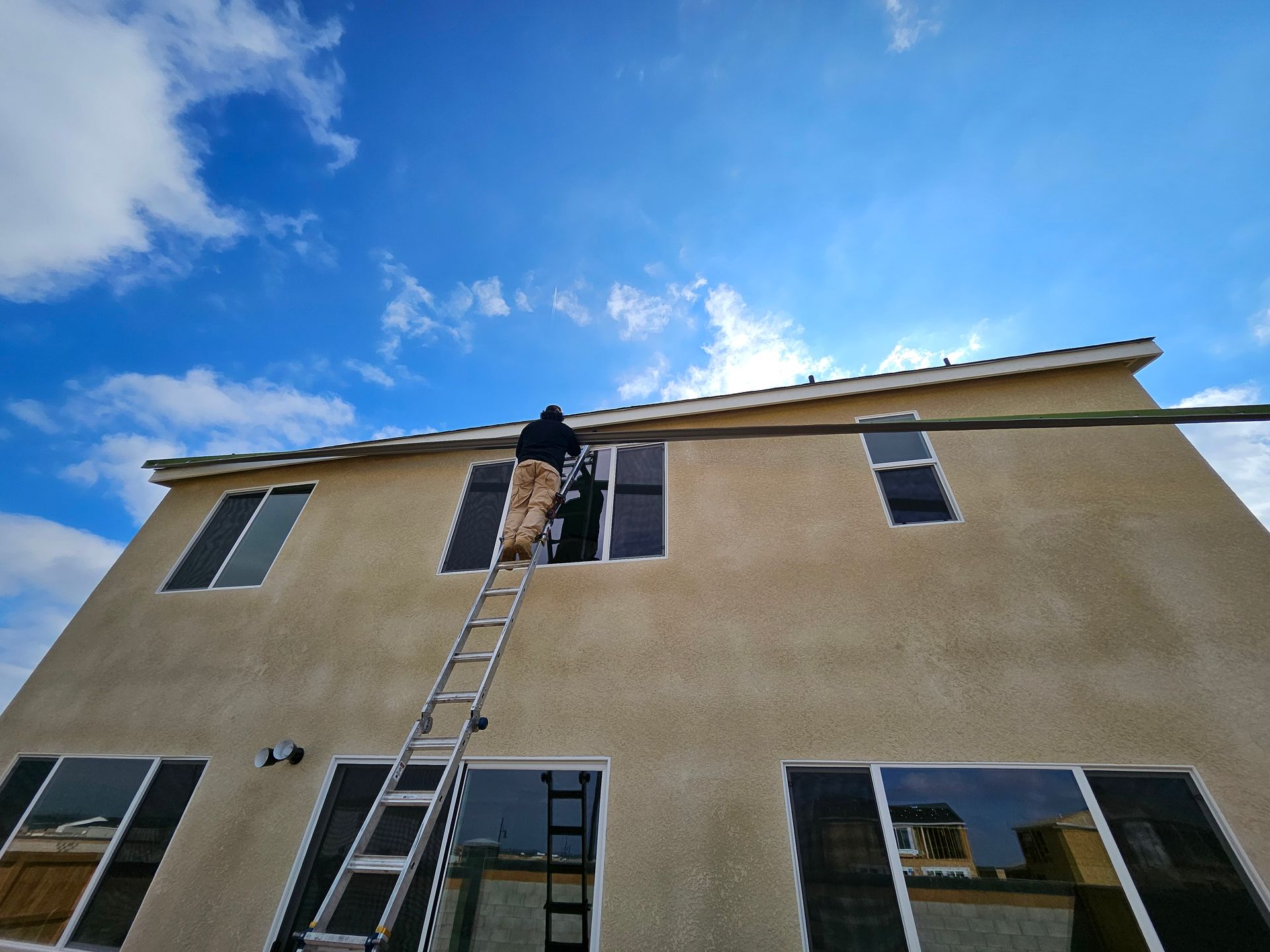 Person on a ladder cleaning windows of a two-story tan house under a blue sky.