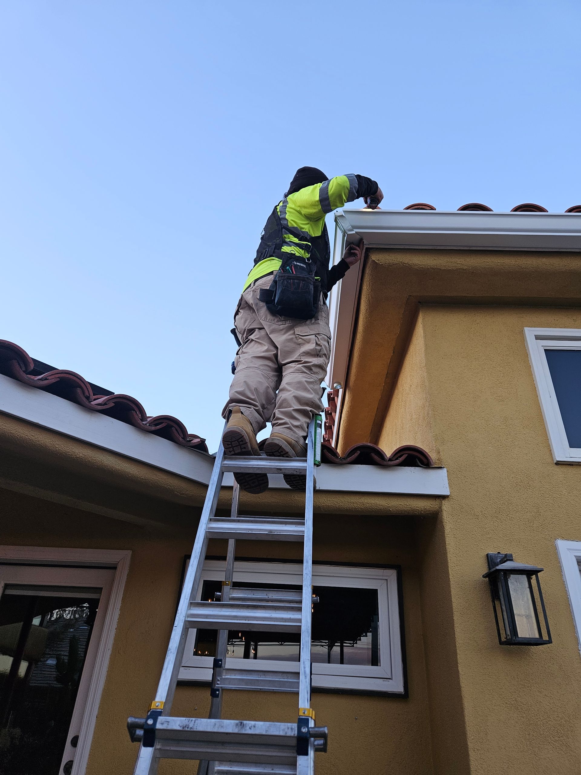 Person on a ladder cleaning a gutter on a yellow stucco house with a red tile roof.