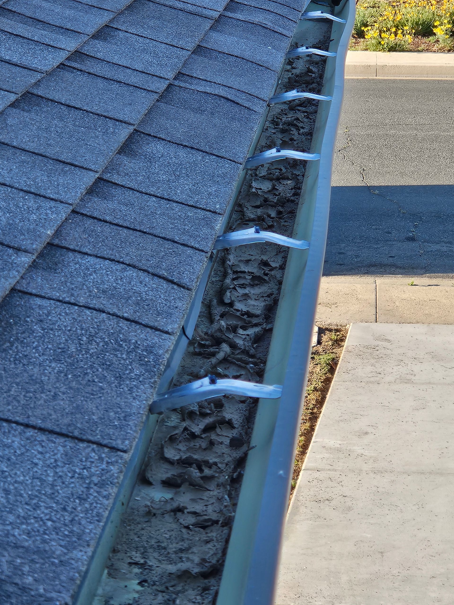A high-angle view of a residential roof gutter heavily clogged with dark, compacted debris and leaves.