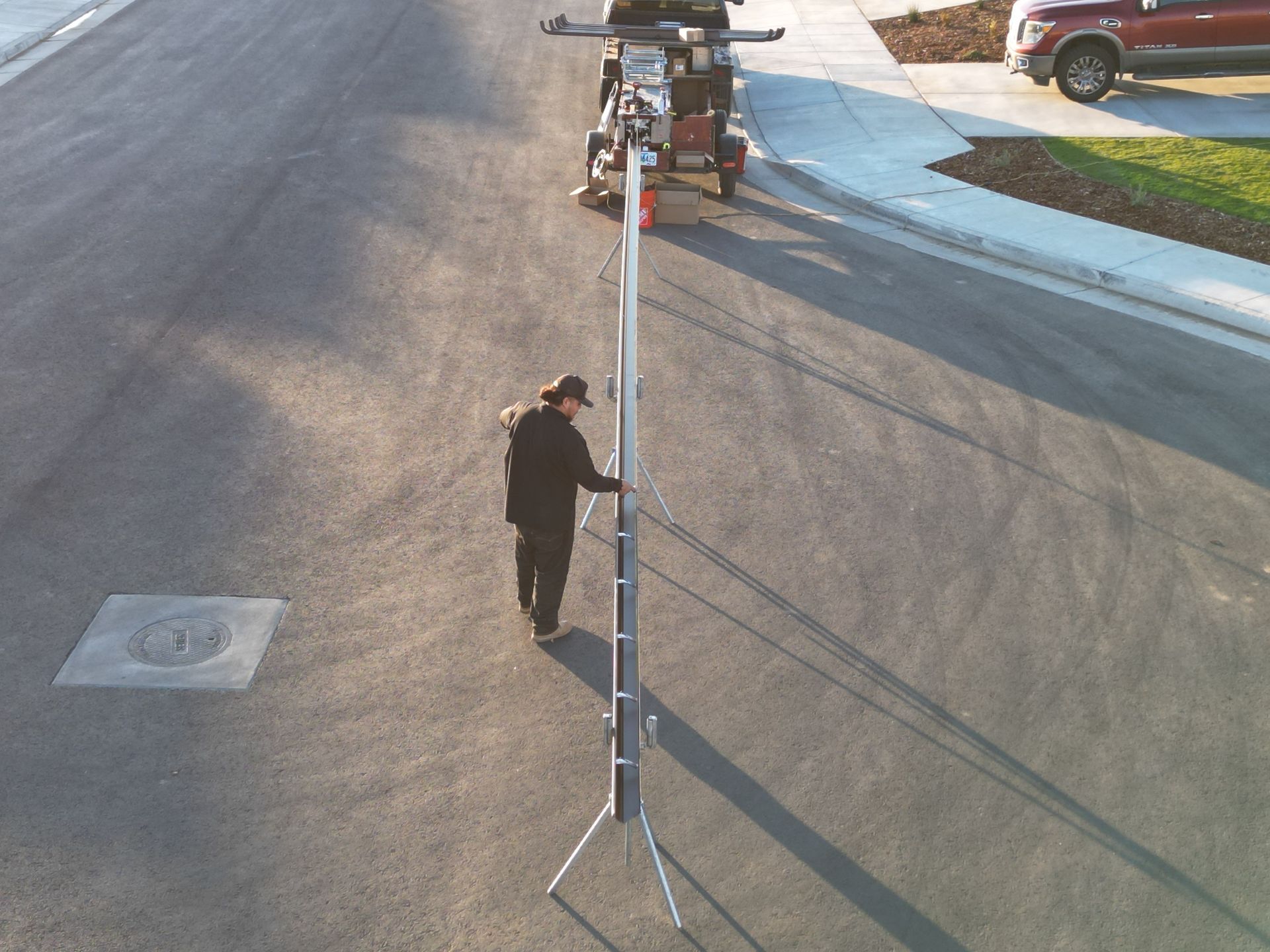 Person setting up a tall light stand on a paved road with equipment nearby and a vehicle in the background.