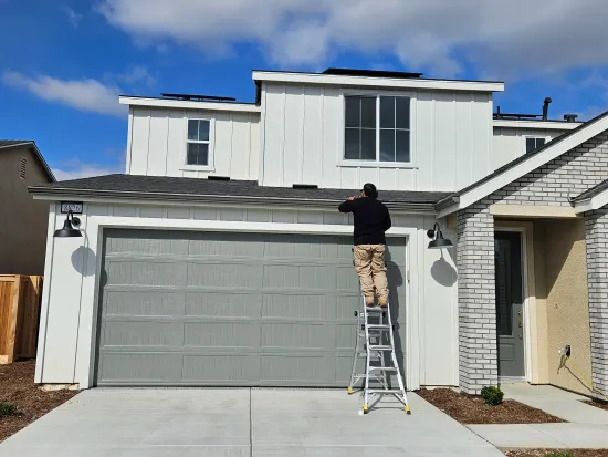 Man on a ladder working on the roof of a white two-story house with a gray garage door.