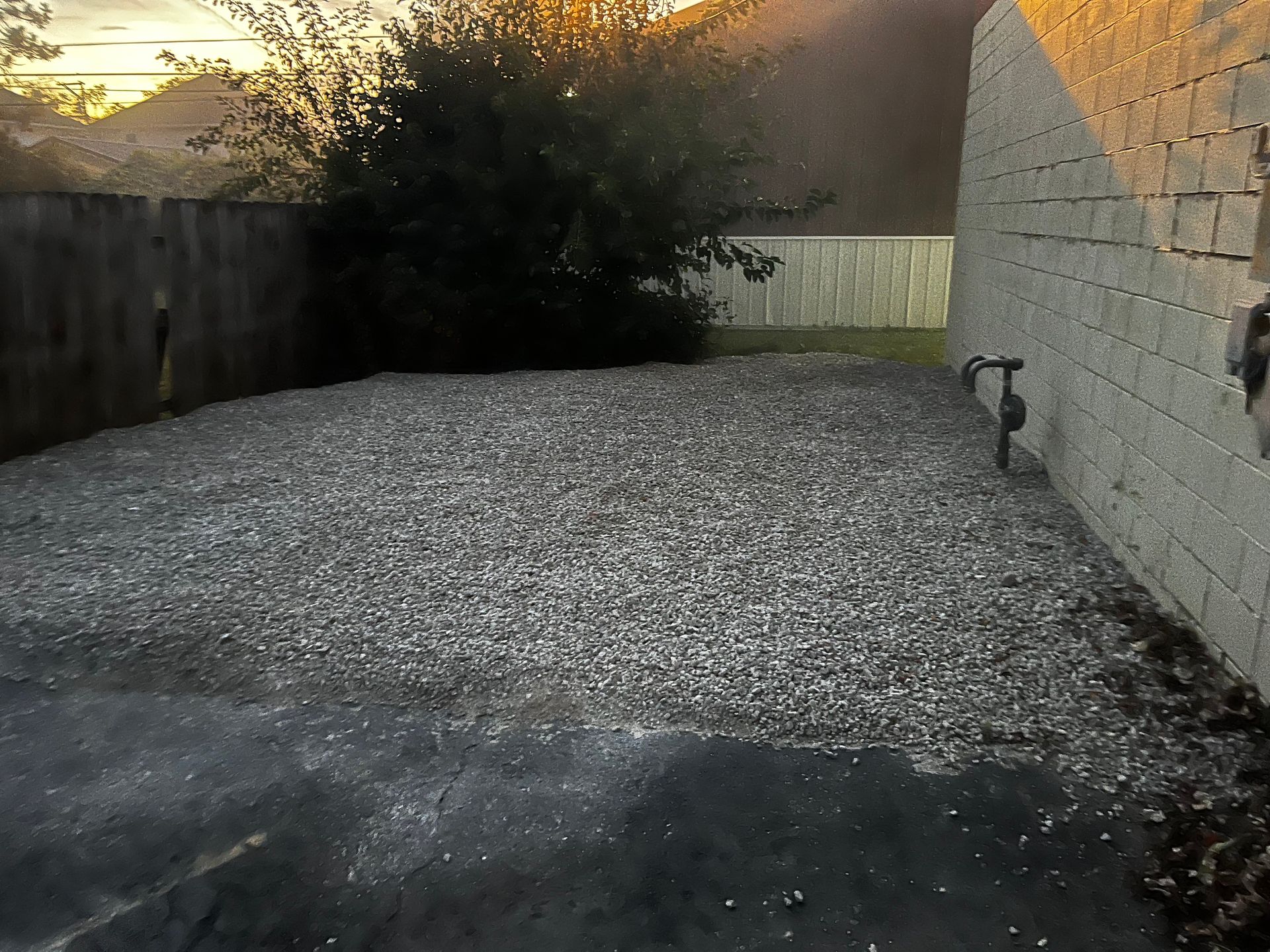 Gravel-covered yard beside a cinder block wall at sunset, with a fence and shrubs in the background