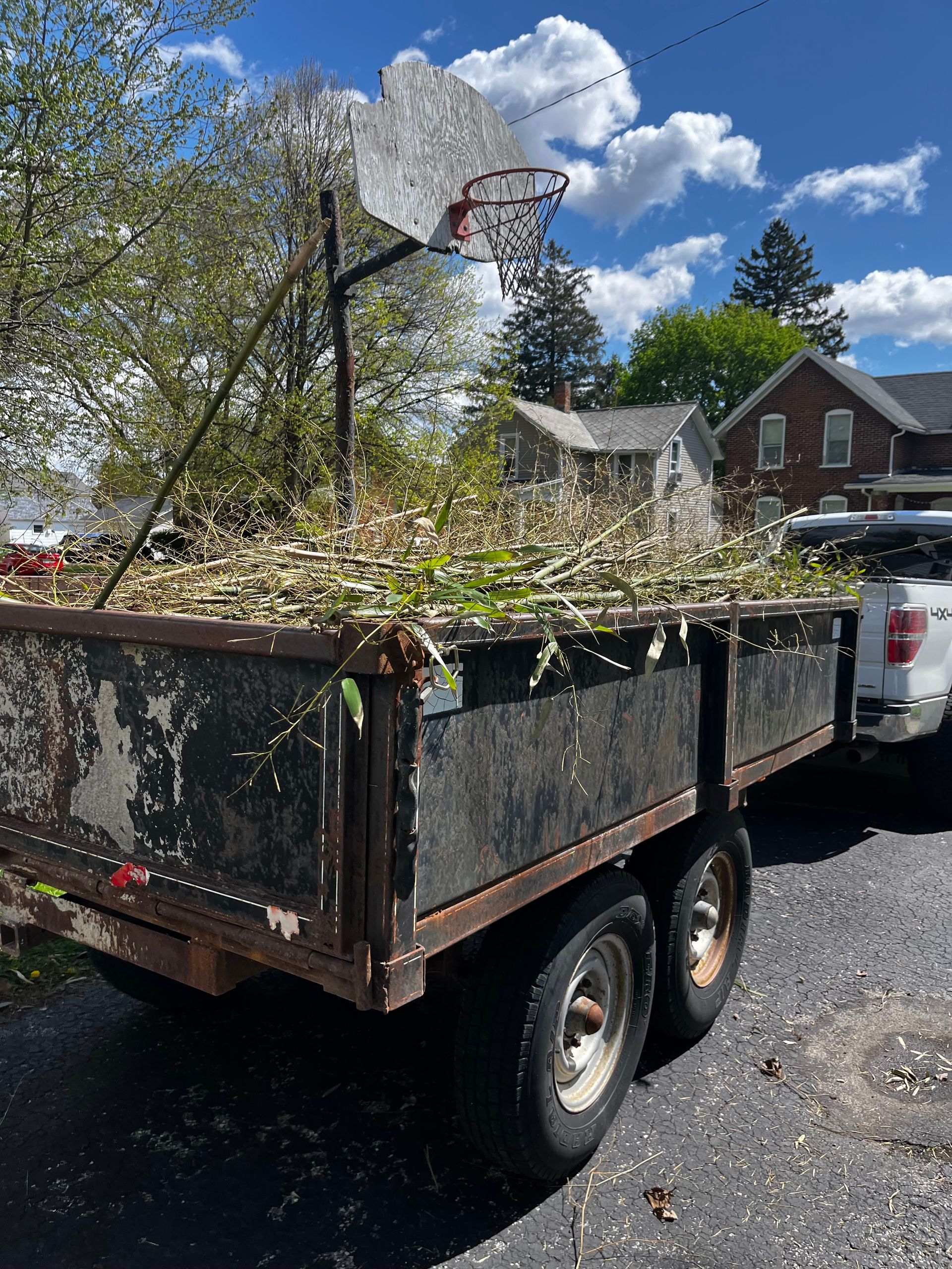 Two-wheel trailer loaded with yard debris and a basketball hoop, parked on a driveway beside a house.