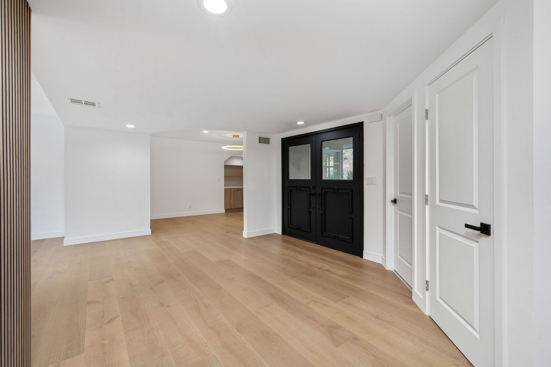 Empty room with light wood flooring, white walls, and black double doors. A hallway and closet are visible in the background.