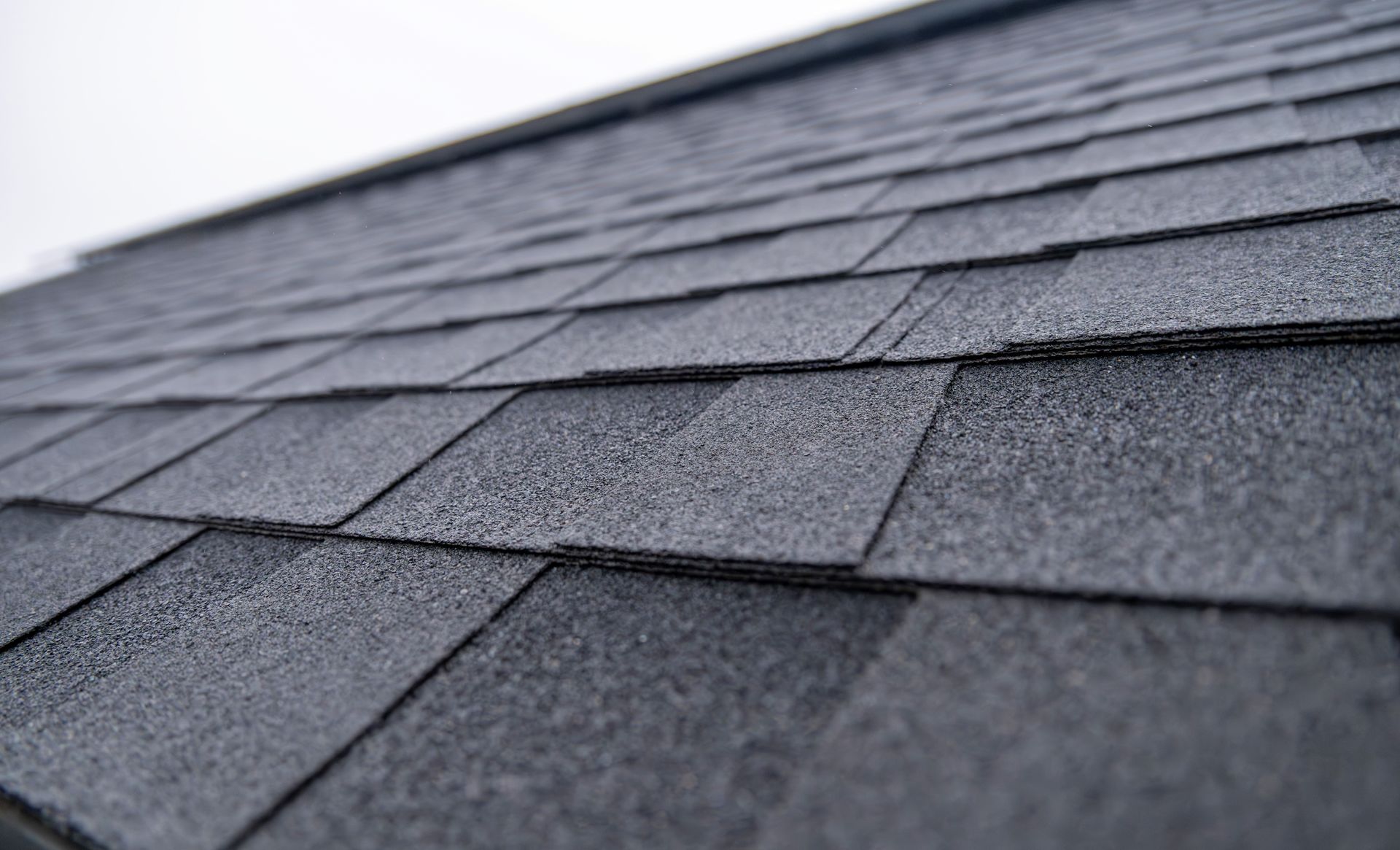 A close-up, angled view of dark grey asphalt roof shingles overlapping in a repeating pattern. A close-up, angled view of dark grey asphalt roof shingles overlapping in a repeating pattern.