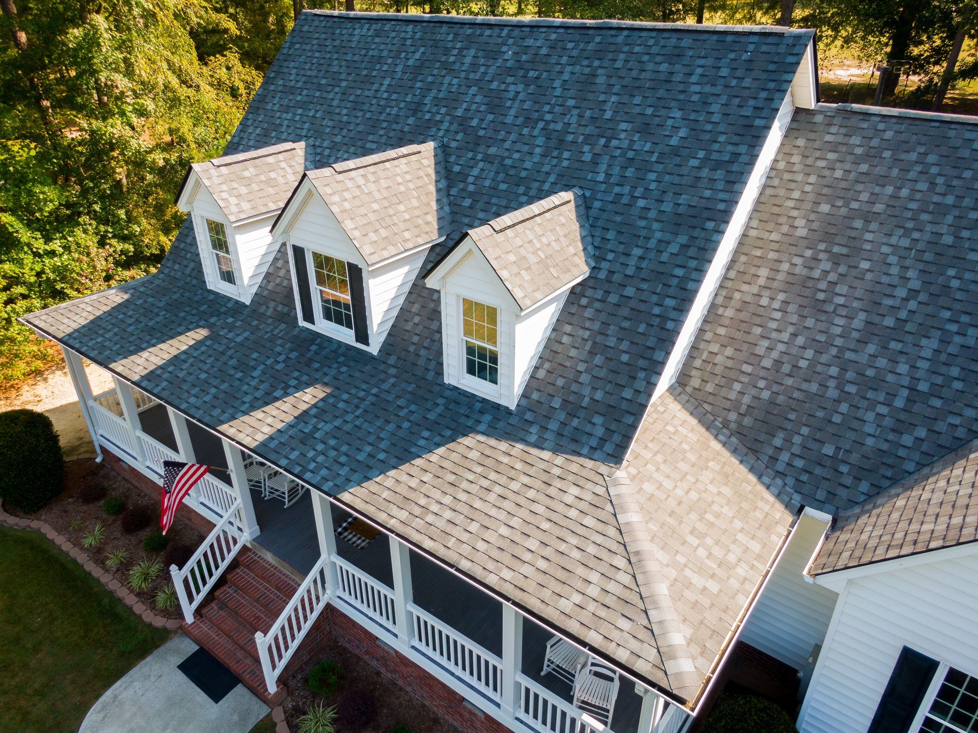 Aerial view of a white house with a multi-gabled dark gray shingled roof and a front porch, surrounded by green trees. Aerial view of a white house with a multi-gabled dark gray shingled roof and a front porch, surrounded by green trees.