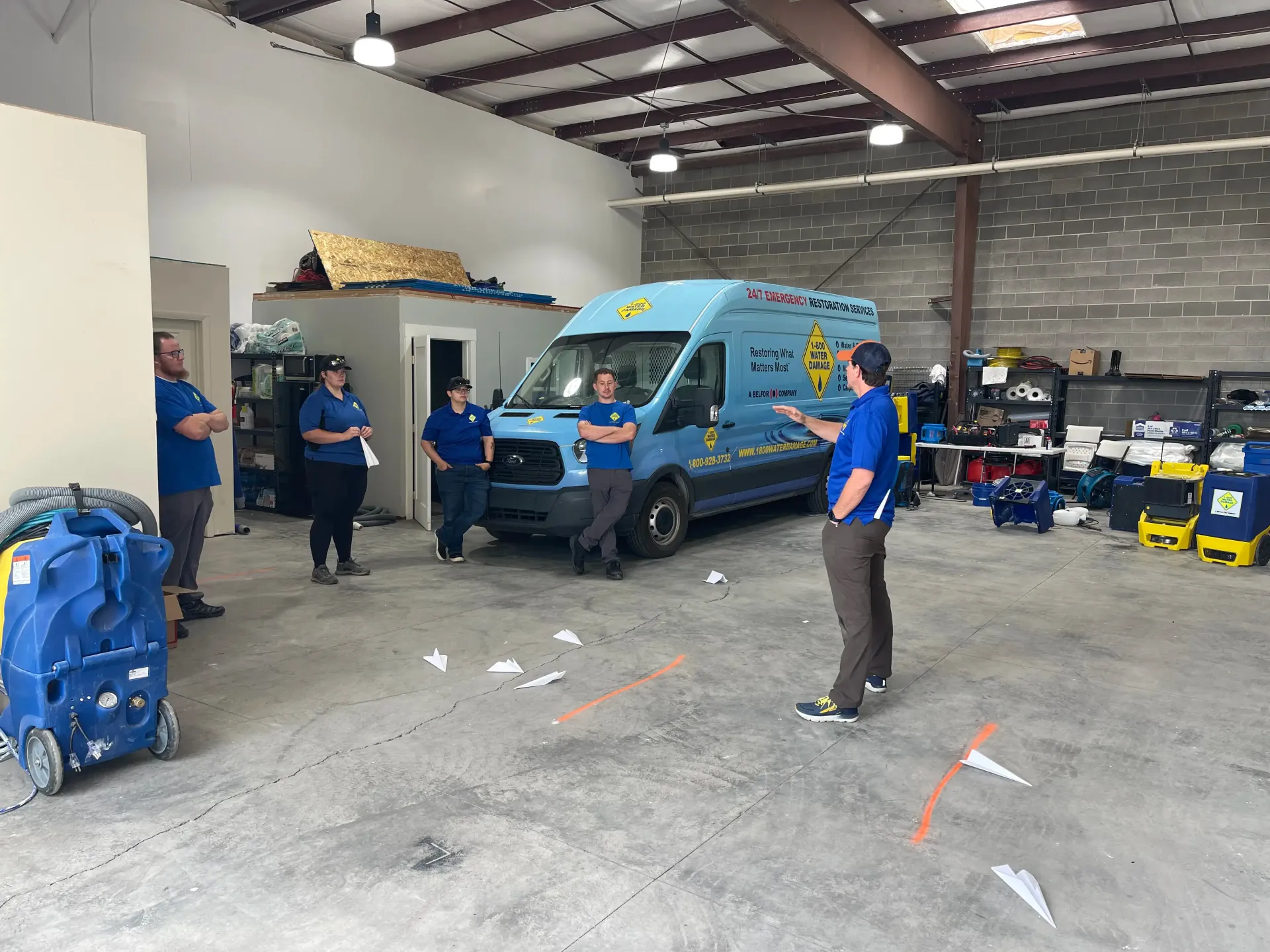 A group of people are standing around a blue van in a warehouse.