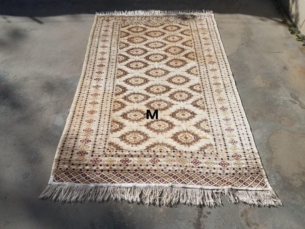 A rectangular area rug with a beige geometric diamond pattern and fringe, laid flat on a paved surface.