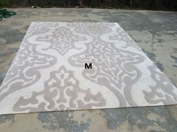 A rectangular, off-white rug featuring a large, repeating light grey damask pattern, laid out on a concrete surface.