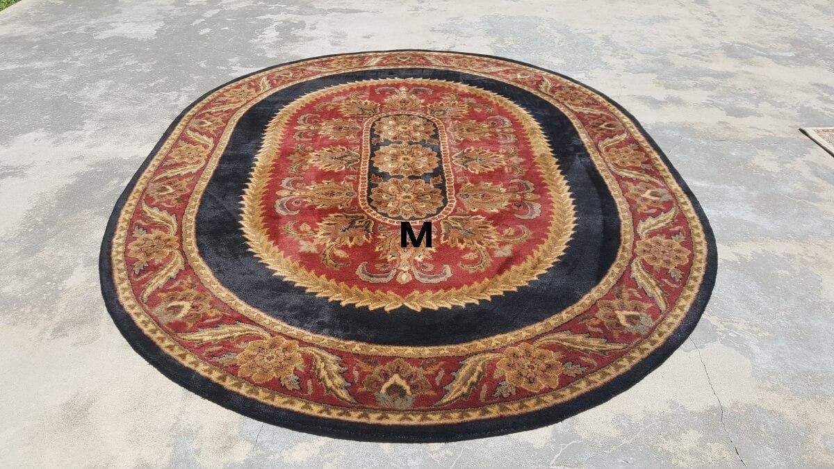 An oval rug with a black center border, red field, and intricate gold, red, and black floral patterns on concrete.