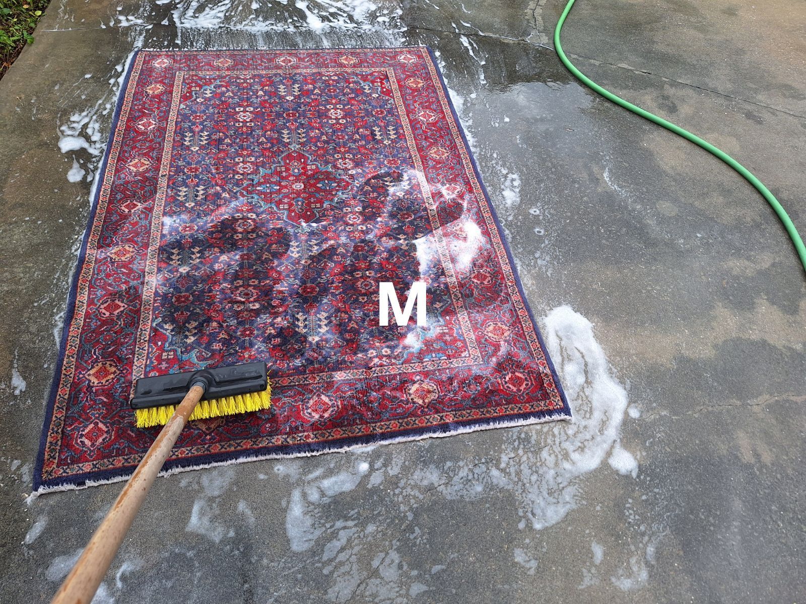 Rug being scrubbed on concrete with a brush, creating suds. Hose on the right.